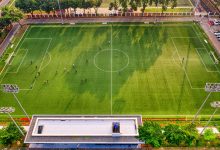 Vista aérea de uma partida de futebol em um estádio de Jacarta, na Indonésia, com jogadores em campo e arquibancadas ao redor.