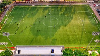 Vista aérea de uma partida de futebol em um estádio de Jacarta, na Indonésia, com jogadores em campo e arquibancadas ao redor.