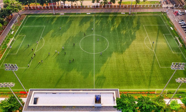 Vista aérea de uma partida de futebol em um estádio de Jacarta, na Indonésia, com jogadores em campo e arquibancadas ao redor.