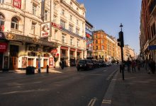 Vista diurna do movimentado distrito teatral do West End, em Londres, com teatros históricos e marcos arquitetônicos.