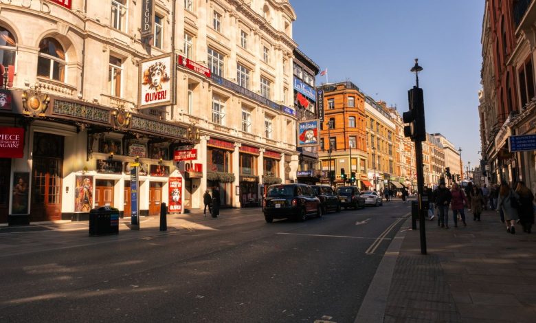 Vista diurna do movimentado distrito teatral do West End, em Londres, com teatros históricos e marcos arquitetônicos.