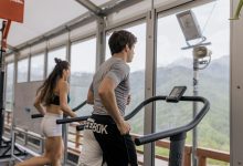 Duas pessoas correm em esteiras lado a lado em uma academia com vista para montanhas.