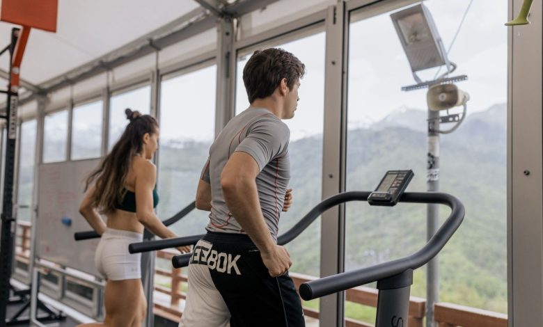 Duas pessoas correm em esteiras lado a lado em uma academia com vista para montanhas.