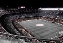 Vista aérea de um grande estádio de futebol contemporâneo, com torcedores nas arquibancadas e o campo iluminado.