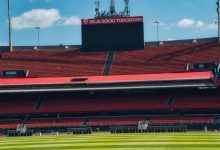 Estádio de futebol vazio, com arquibancadas vermelhas, placar e campo gramado sob céu claro.