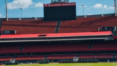 Estádio de futebol vazio, com arquibancadas vermelhas, placar e campo gramado sob céu claro.