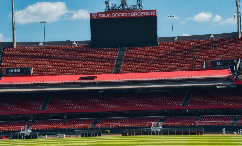 Estádio de futebol vazio, com arquibancadas vermelhas, placar e campo gramado sob céu claro.